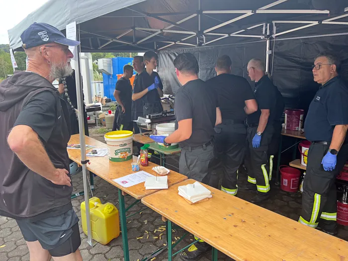 Männer in dunklen Uniformen bereiten unter einem Zelt Verpflegung an Holztischen vor.