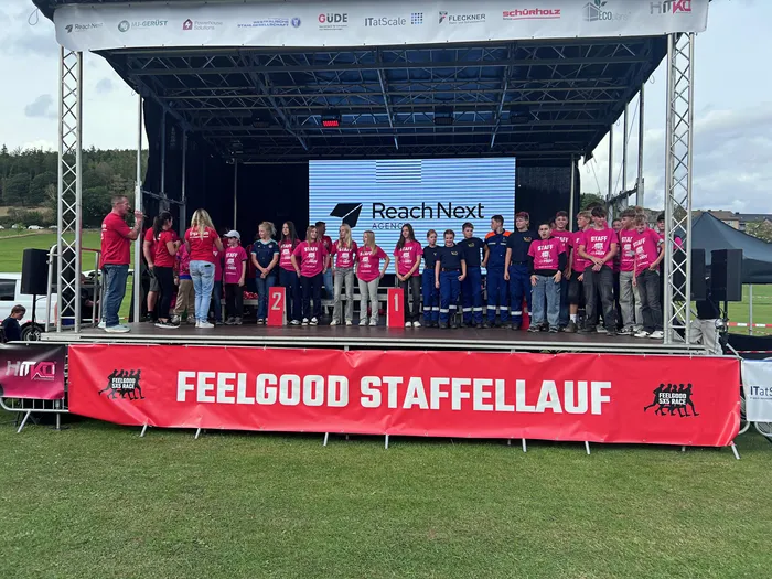 Eine Gruppe von Menschen in pinkfarbenen T-Shirts steht auf einer Bühne vor einem großen Bildschirm. Vor der Bühne ist ein rotes Banner mit der Aufschrift „FEELGOOD STAFFELLAUF“ gespannt.