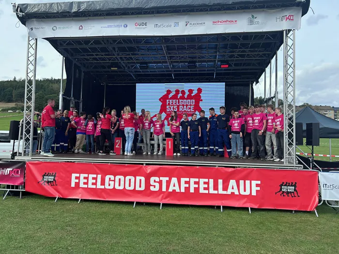 Eine Gruppe von Menschen posiert auf einer Bühne vor einer Leinwand und einem Banner mit der Aufschrift „FEELGOOD STAFFELLAUF“.