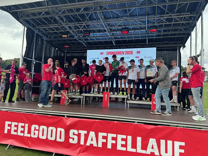Teilnehmer des Feelgood-Staffellaufs stehen mit roten Taschen auf einer Bühne. Ein Banner mit der Aufschrift „FEELGOOD STAFFELLAUF“ ist im Vordergrund zu sehen.