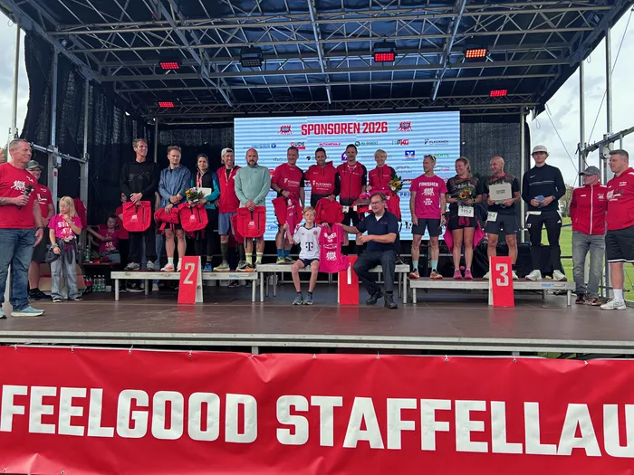 Teilnehmer des Feelgood-Staffellaufs stehen auf einer Bühne vor einer Sponsorenwand und halten Trophäen sowie rote Taschen.