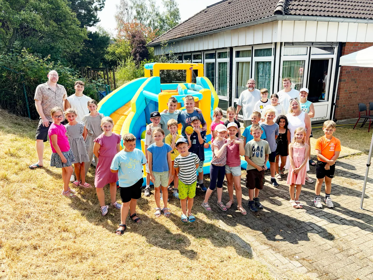 Eine Gruppe von Kindern und Erwachsenen posiert vor einer blau-gelben aufblasbaren Rutsche im Freien.