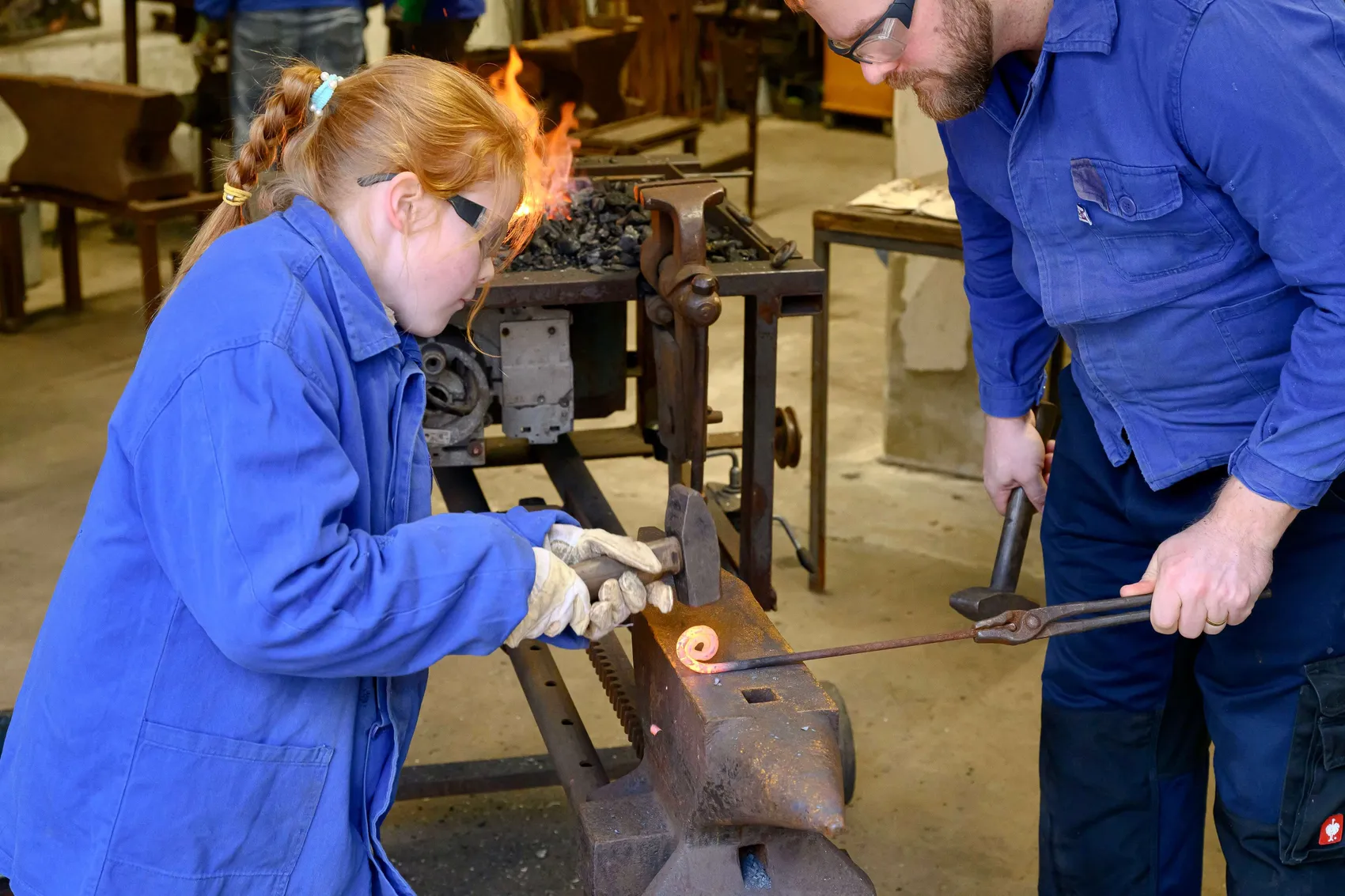 Eine junge Person mit roten Haaren und ein Mann in blauen Arbeitsanzügen bearbeiten mit Hammer und Zange ein Metallstück auf einem Amboss.