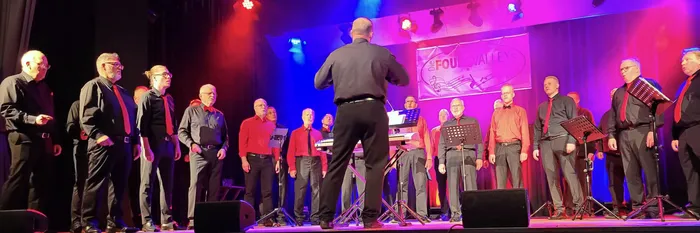 Ein A-cappella-Ensemble singt auf einer Bühne unter farbiger Beleuchtung, geleitet von einem Dirigenten. Im Hintergrund hängt ein Banner mit „the FOUR VALLEYS“.
