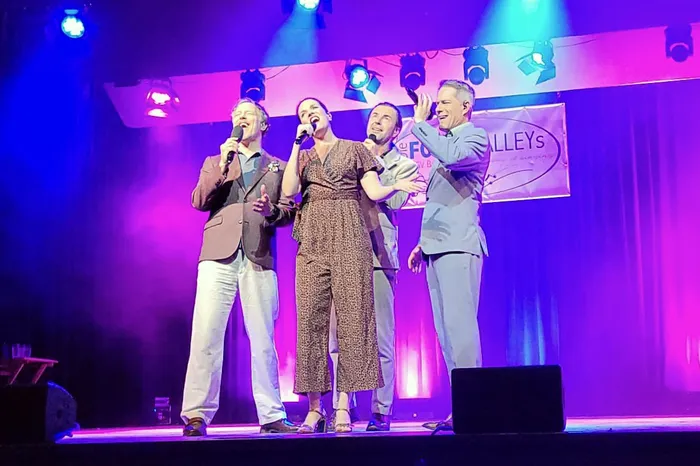 Vier Sänger unter violettem Bühnenlicht singen in Mikrofone vor einem „Four Valleys“-Banner.