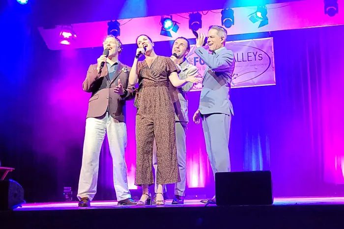 Vier Mitglieder des A-cappella-Ensembles LaLeLu singen mit Mikrofonen auf einer Bühne unter lila und blauer Beleuchtung.