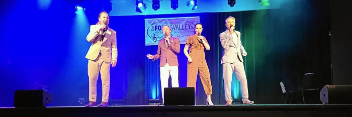 Vier Sänger stehen mit Mikrofonen auf einer Bühne unter blauem und grünem Licht vor einem Banner der Gruppe „Four Valleys“.