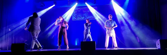 Vier Sänger stehen unter blauem Bühnenlicht auf einer Bühne und singen in Mikrofone. Im Hintergrund hängt ein Banner der Gruppe „The Four Valleys“.