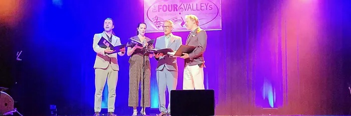 Vier Sänger stehen unter violettem Bühnenlicht und singen mit Musikmappen vor einem Banner der Gruppe 'Four Valleys'.
