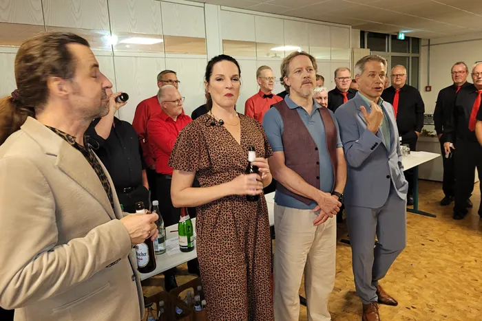 Mitglieder des A-cappella-Ensembles LaLeLu stehen in einer Gruppe in einem Raum, ein Mann im hellblauen Anzug gestikuliert beim Sprechen.