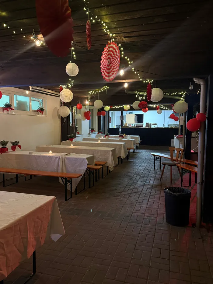 Der Festsaal ist für eine Feier dekoriert: Lange Tische mit weißen Tischdecken stehen bereit, darüber hängen Lichterketten und rote Papierlaternen. Im Hintergrund ist ein Tresen zu sehen, der Raum ist mit roten Dekorationen geschmückt.