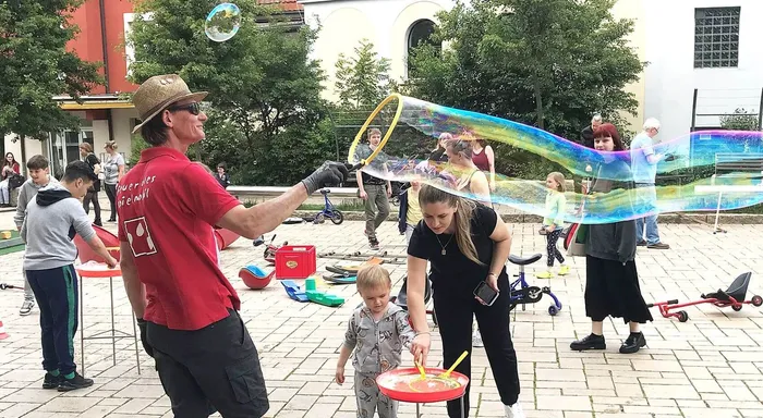 Ein Mann mit Strohhut macht mit Kindern Seifenblasen im Rosengarten. Im Hintergrund stehen weitere Menschen und Spielgeräte des 
