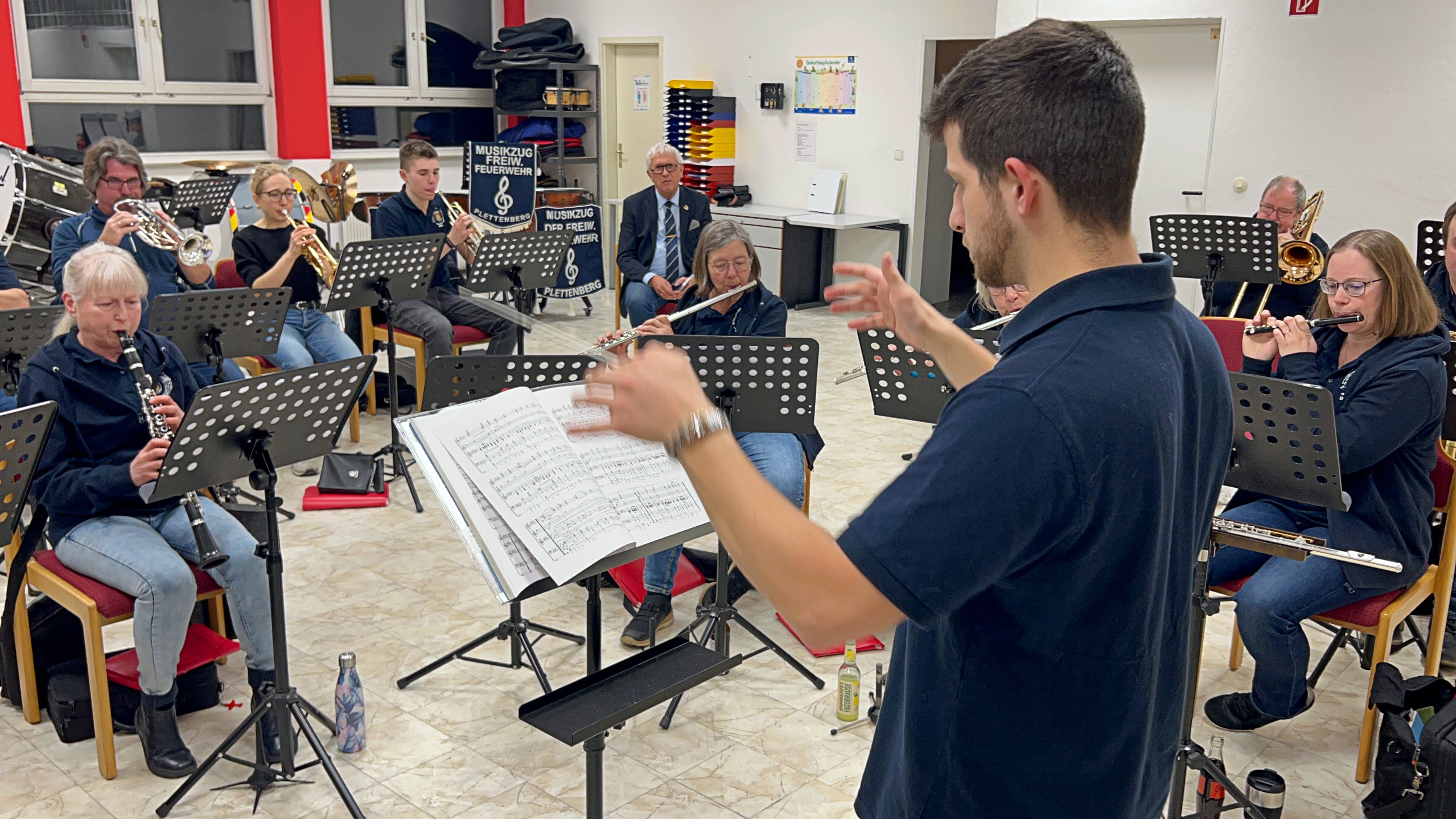 Ein Dirigent steht mit dem Rücken zum Betrachter vor einem Orchester, das verschiedene Blasinstrumente spielt. Im Hintergrund Banner mit der Aufschrift „Musikzug der Freiwilligen Feuerwehr Plettenberg“.