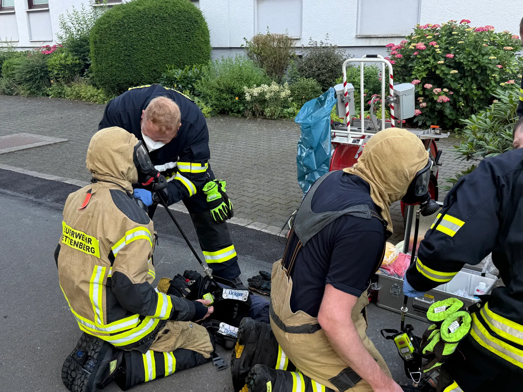 Feuerwehrleute in Schutzkleidung und mit Atemschutzgeräten versorgen eine Person, die auf dem Boden sitzt. Im Hintergrund steht ein Rettungswagen.