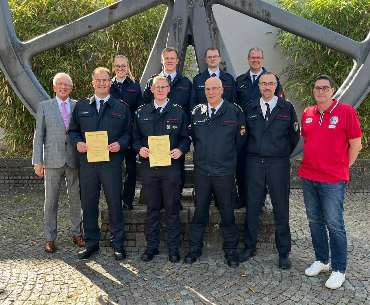 Feuerwehrleute in Uniform und Bürgermeister Ralf Beßler posieren mit Urkunden vor einem großen Metallrad.