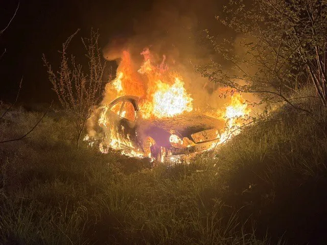 Ein brennendes Auto steht in Flammen, umgeben von trockenem Gras und Büschen in der Dunkelheit.