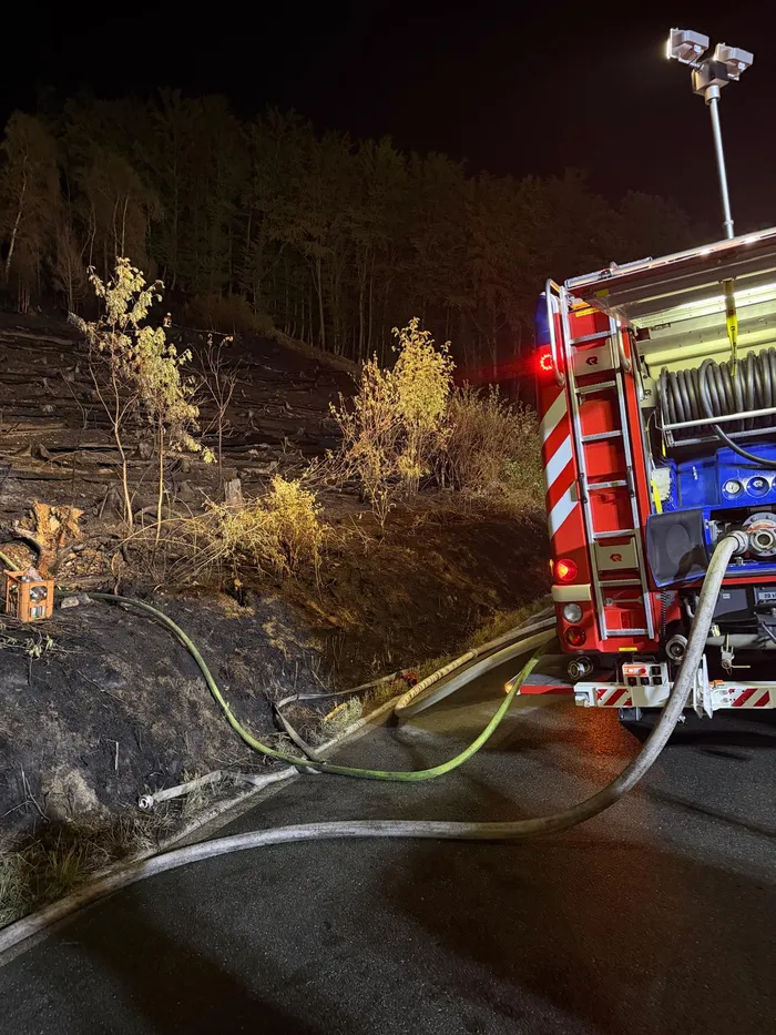 Feuerwehrfahrzeug mit Schläuchen, die zu einem verkohlten Waldhang führen, bei nächtlichen Löscharbeiten.