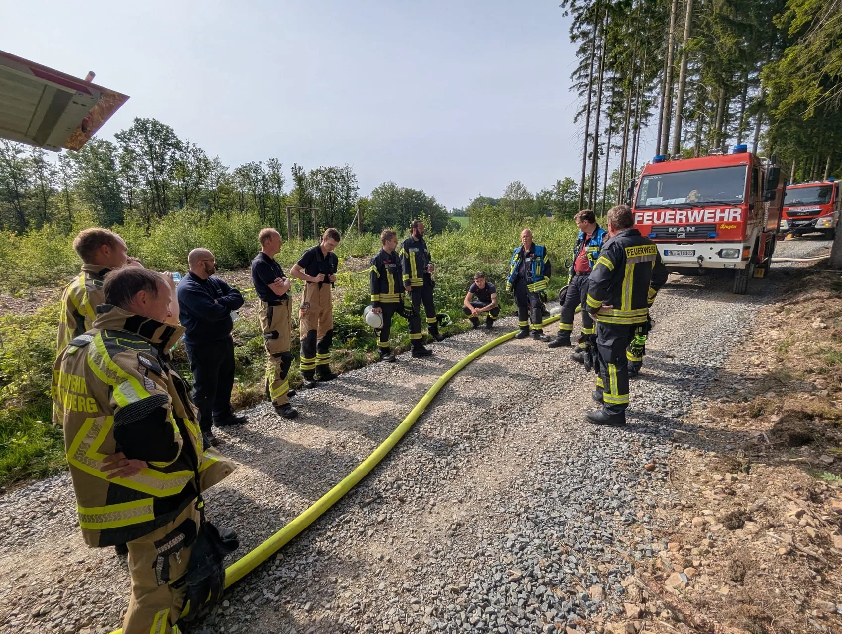Eine Gruppe von Feuerwehrleuten in voller Montur steht auf einem Schotterweg zusammen und bespricht sich. Im Hintergrund steht ein rotes Feuerwehrauto mit der Aufschrift „Feuerwehr“.