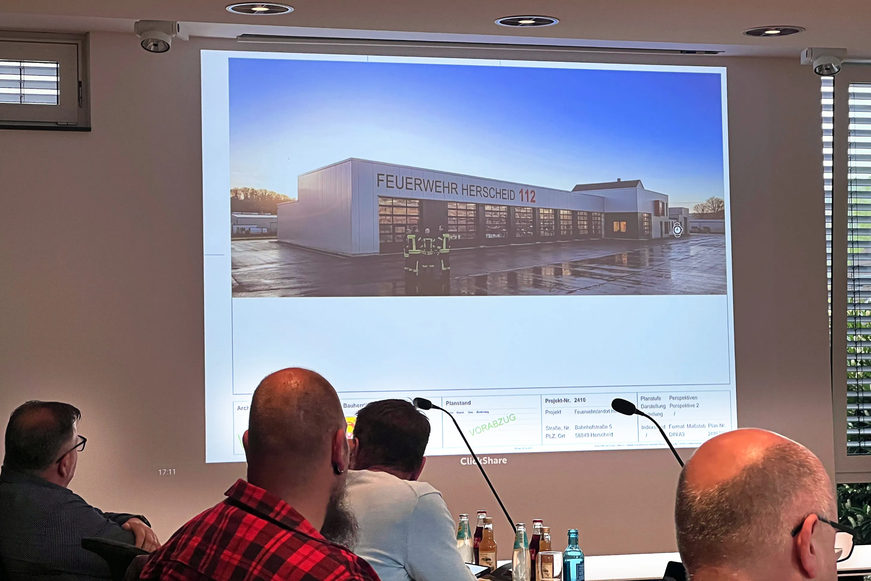 Architektonische Visualisierung einer modernen Fahrzeughalle für die Feuerwehr Herscheid, die auf einer Präsentation im Hintergrund eines Besprechungsraums zu sehen ist. Im Vordergrund sitzen drei Männer an einem Tisch mit Mikrofonen.