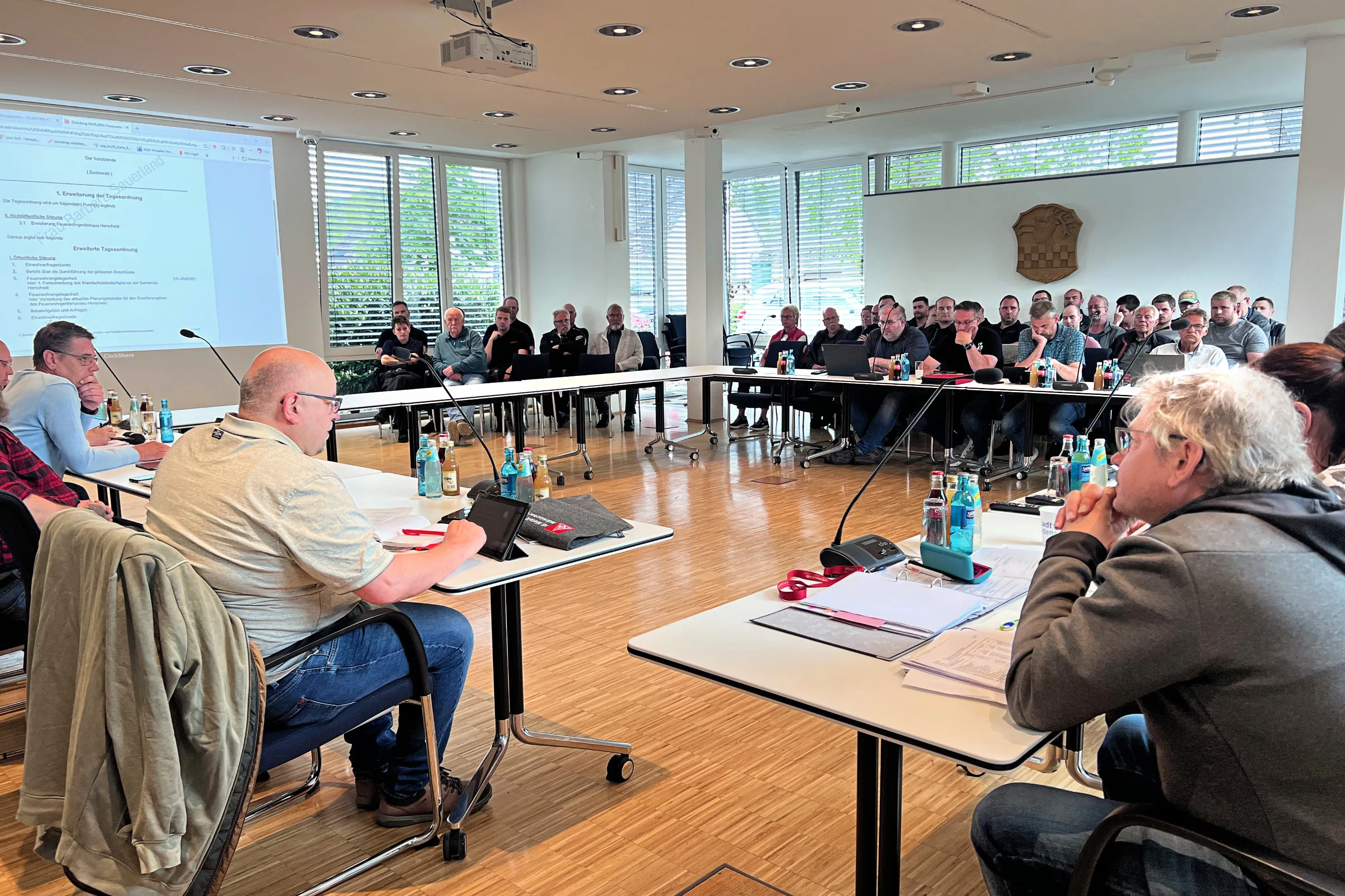 Versammlung von Feuerwehrleuten in einem hellen Besprechungsraum, die einem Vortrag auf einer Präsentation folgen. Einige sitzen an Tischen, andere auf Stühlen in einem Halbkreis.