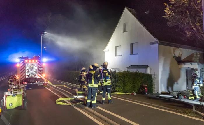 Feuerwehrleute in gelben Schutzanzügen stehen vor einem zweigeschossigen Gebäude, aus dessen Dach Rauch quillt. Im Hintergrund ein rotes Feuerwehrauto mit Blaulicht.