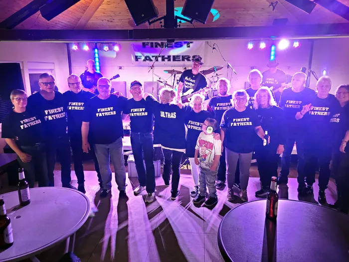 Die Hardrockcoverband Finest Fathers posiert mit einer Gruppe von Menschen auf einer Bühne vor einem Band-Banner unter lila Bühnenlicht.