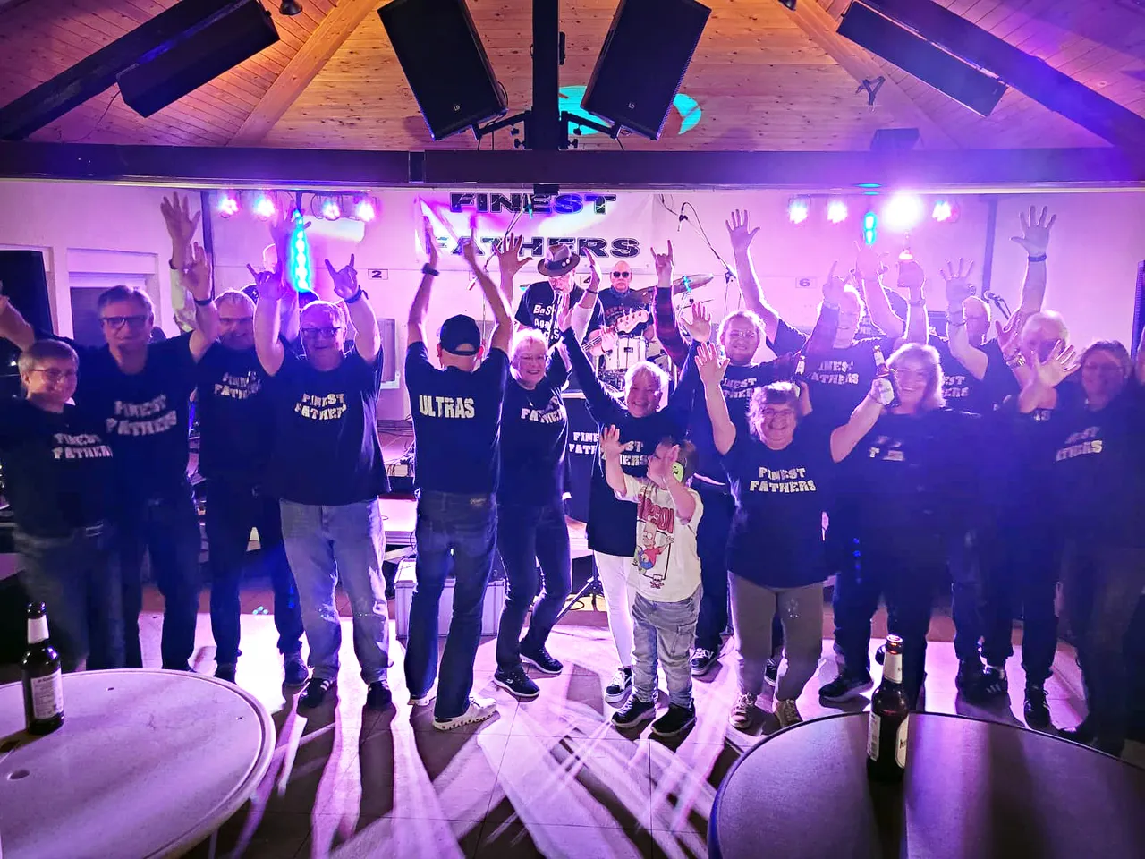 Eine Gruppe von Menschen in „Finest Fathers“-T-Shirts posiert mit erhobenen Händen unter bunter Bühnenbeleuchtung.