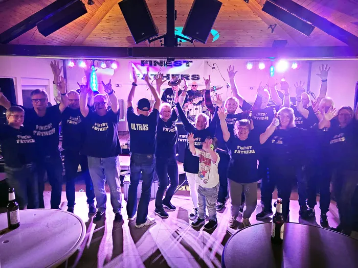 Eine Gruppe von Menschen in „Finest Fathers“-T-Shirts posiert mit erhobenen Händen unter bunter Bühnenbeleuchtung.