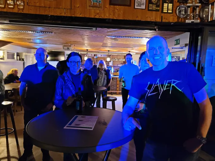 Menschen stehen in einer Bar bei blauem Licht um einen Tisch, im Vordergrund ein lächelnder Mann in einem schwarzen „Slayer“-T-Shirt.