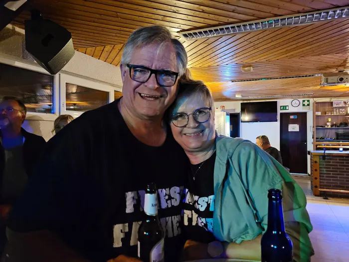 Ein lächelndes Paar posiert eng beieinander; der Mann trägt ein schwarzes T-Shirt der Band Finest Fathers.