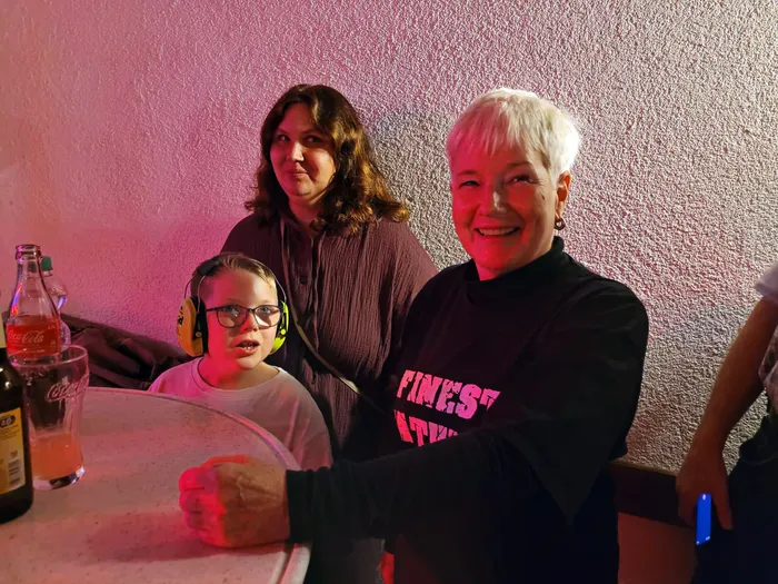 Eine lächelnde Frau im „Finest Fathers“-Shirt, eine Frau und ein Junge mit gelbem Gehörschutz stehen an einem Tisch mit Getränken.