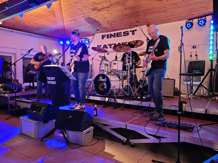 Die Hardrockcoverband Finest Fathers spielt auf einer Bühne mit farbiger Beleuchtung und einem Band-Banner im Hintergrund.