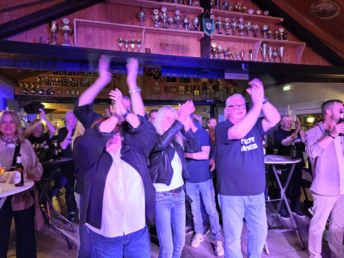 Menschen tanzen und jubeln mit erhobenen Händen in einem holzgetäfelten Raum mit zahlreichen Trophäen an den Wänden.