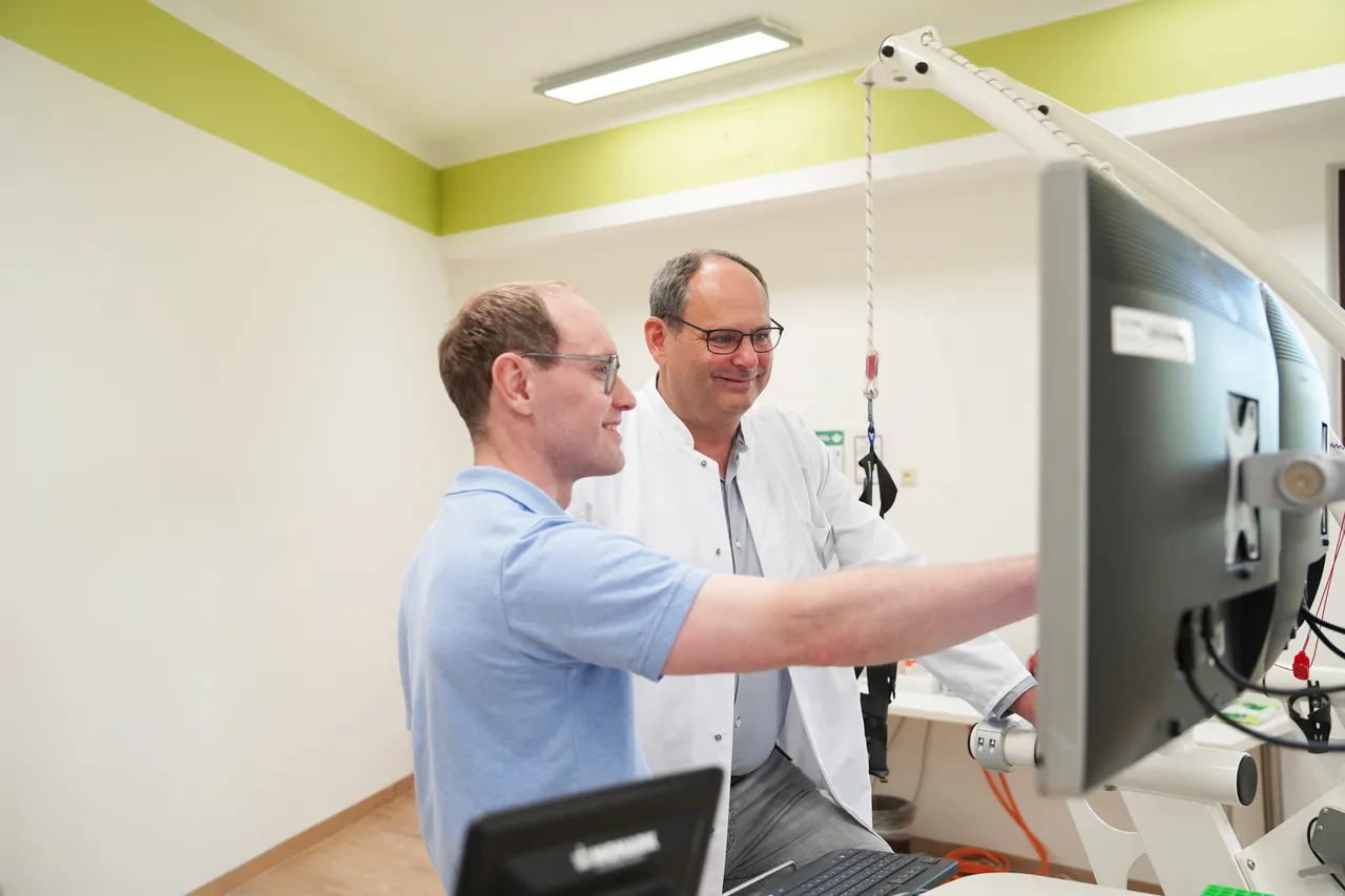 Zwei Männer betrachten gemeinsam einen Computerbildschirm. Der Mann links trägt ein hellblaues Hemd, der Mann rechts einen weißen Kittel und eine Brille.