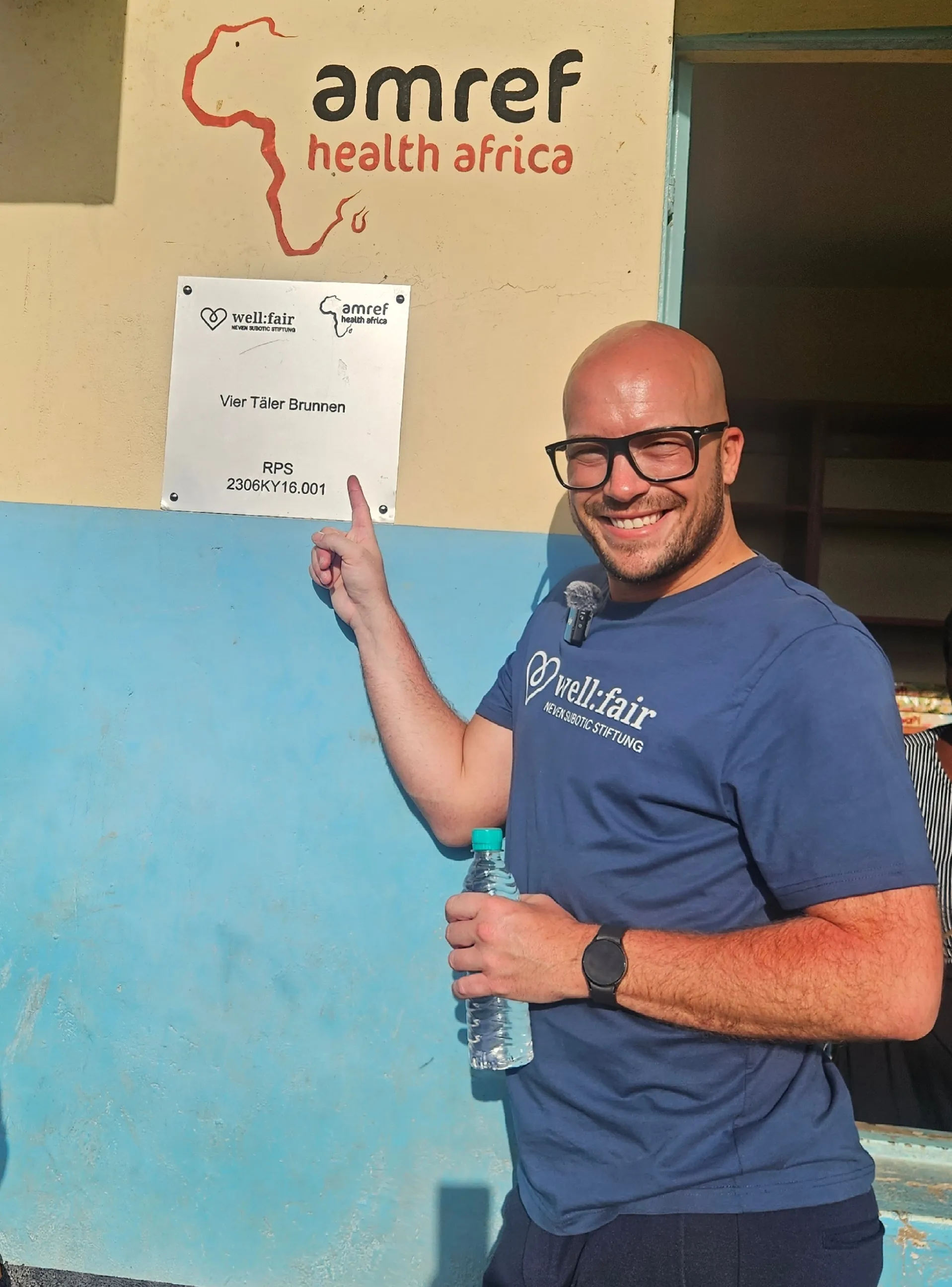 Mann mit Bart und Brille hält Mikrofon vor einer blauen Wand mit Logos von amref health africa und well:fair Stiftung. Im Hintergrund ein Schild mit der Aufschrift „Vier Taler Brunnen“.