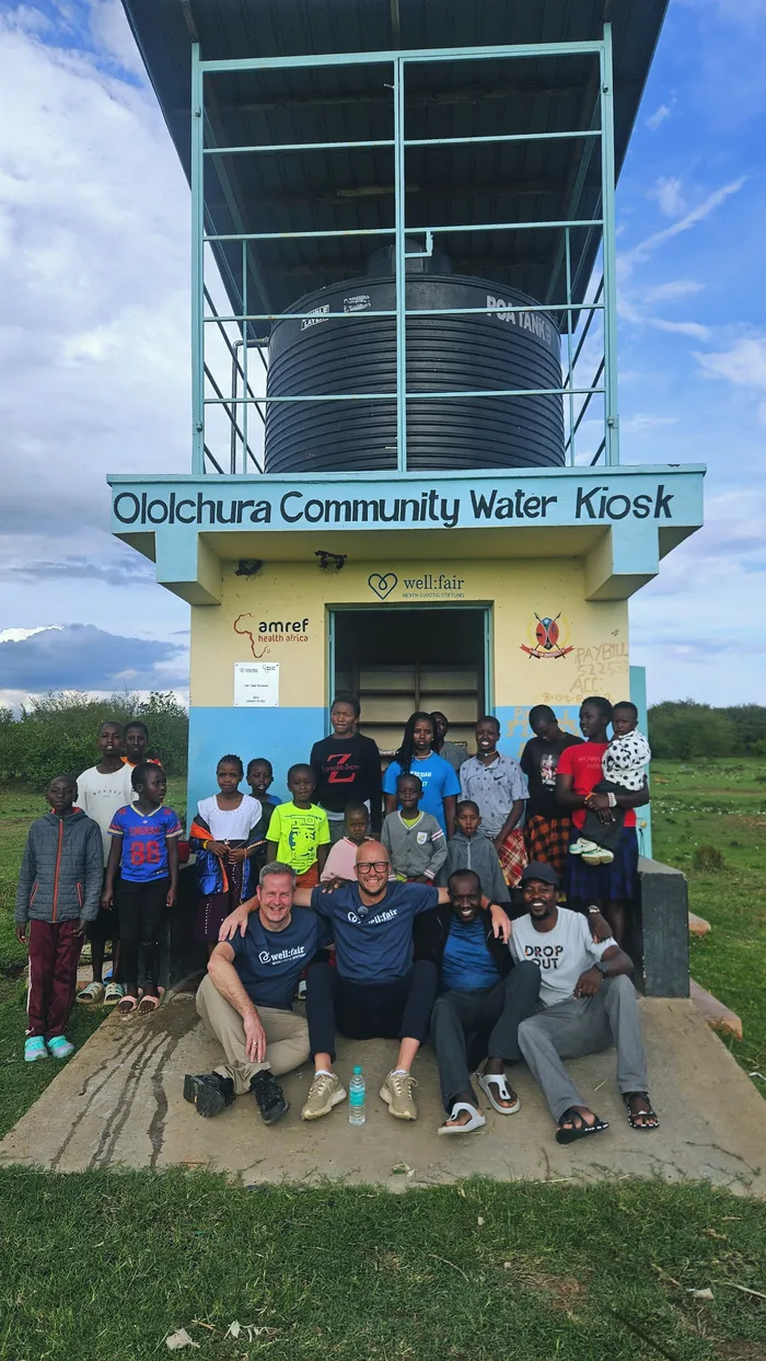 Eine Gruppe von etwa 30 Menschen, darunter Kinder und Männer, posiert vor einem Wassertank mit Solaranlage und dem Schriftzug „Ololchura Community Water Kiosk“. Zwei europäisch aussehende Männer sitzen im Vordergrund.