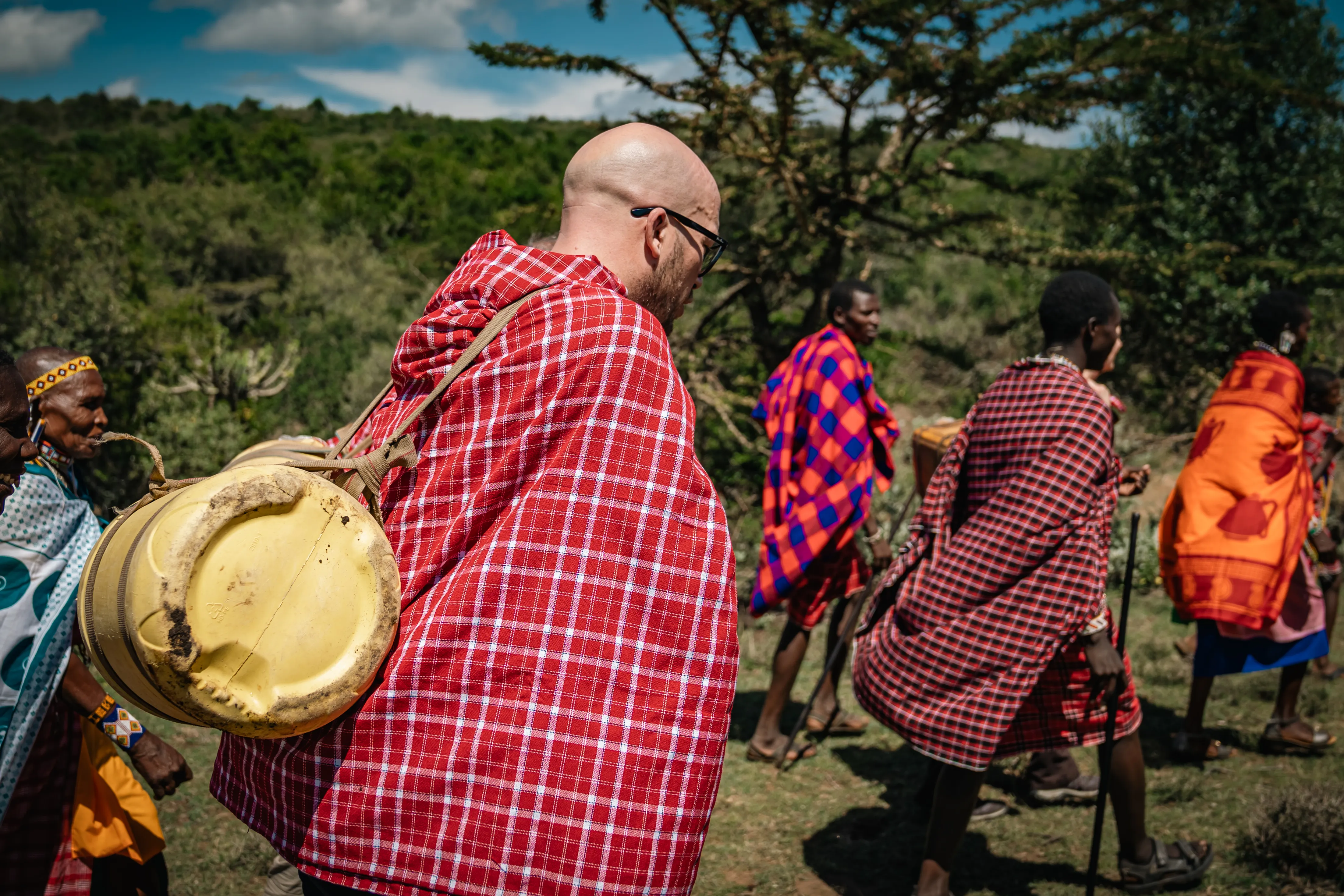 Ein Mann mit Glatze und Bart geht mit einem großen gelben Kanister auf dem Rücken vor einer Gruppe von Menschen in traditioneller Kleidung durch eine Landschaft in Kenia.
