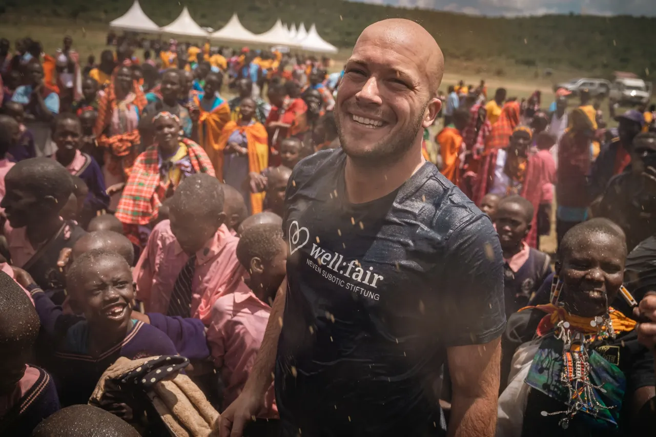 Ein lächelnder Mann mit Glatze und T-Shirt der „well:fair“ Stiftung steht inmitten einer Gruppe von Kindern und Erwachsenen in Kenia. Im Hintergrund sind weitere Menschen und eine Landschaft zu sehen.