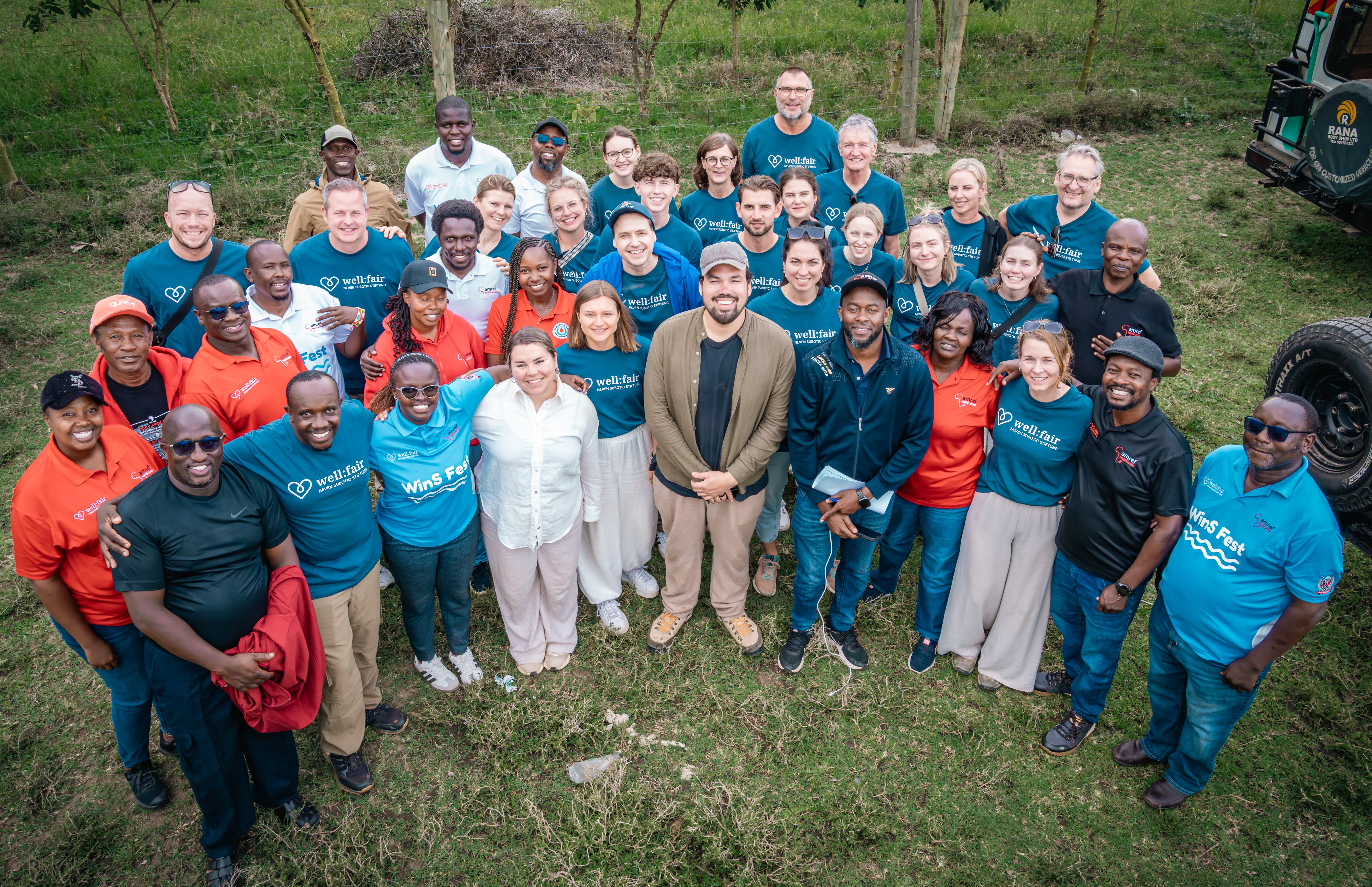 Eine Gruppe von etwa 30 Personen steht lächelnd für ein Gruppenfoto zusammen. Viele tragen T-Shirts mit dem Aufdruck „well:fair“, andere orangefarbene Poloshirts. Im Zentrum steht ein Mann in einem dunklen Jackett und hellen Hosen.