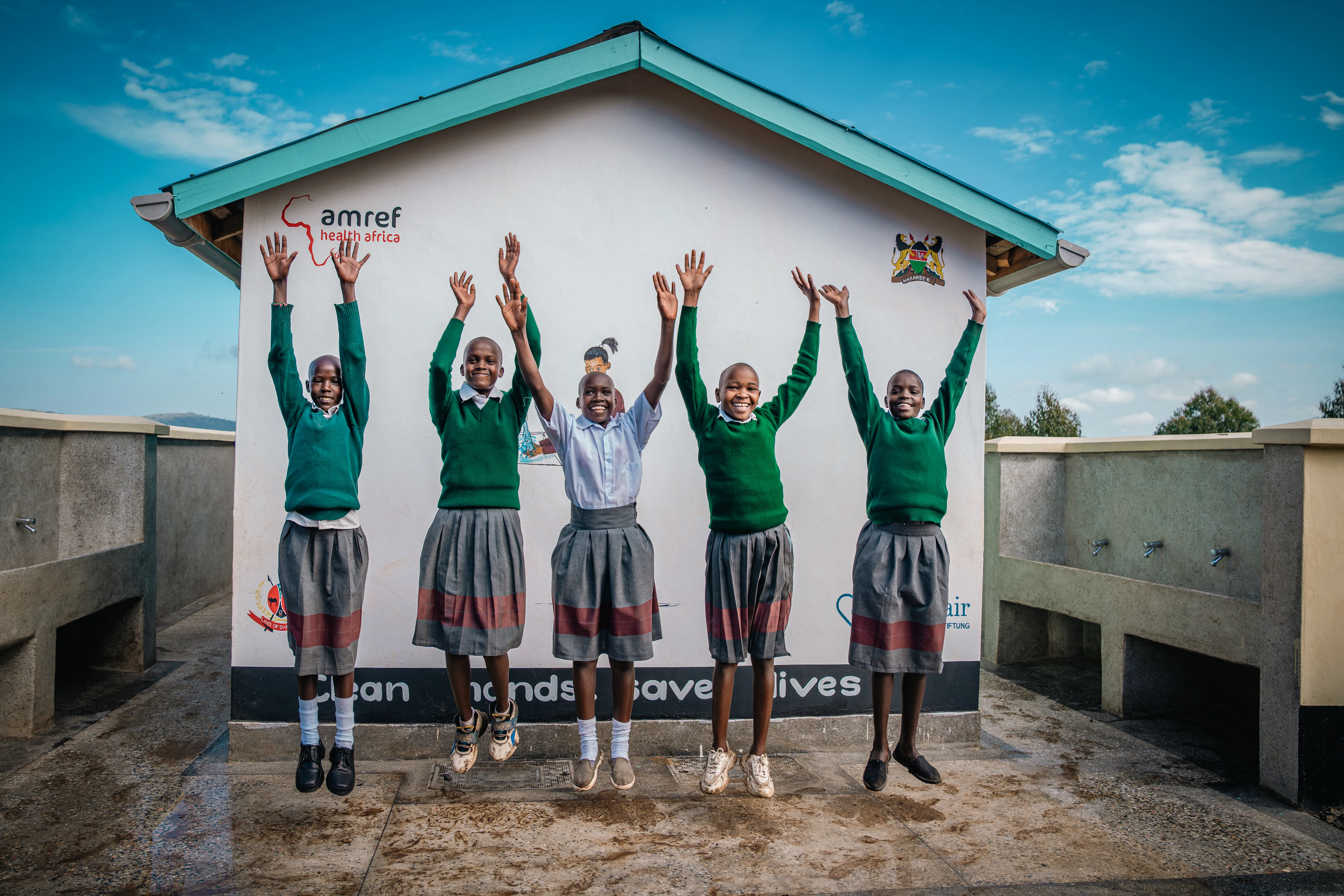 Fünf Kinder in Schuluniformen stehen vor einem Gebäude mit dem Aufdruck „Amref Health Africa“ und heben die Arme in die Luft. Das Gebäude dient als Hygieneeinrichtung mit dem Slogan „Clean hands save lives“.