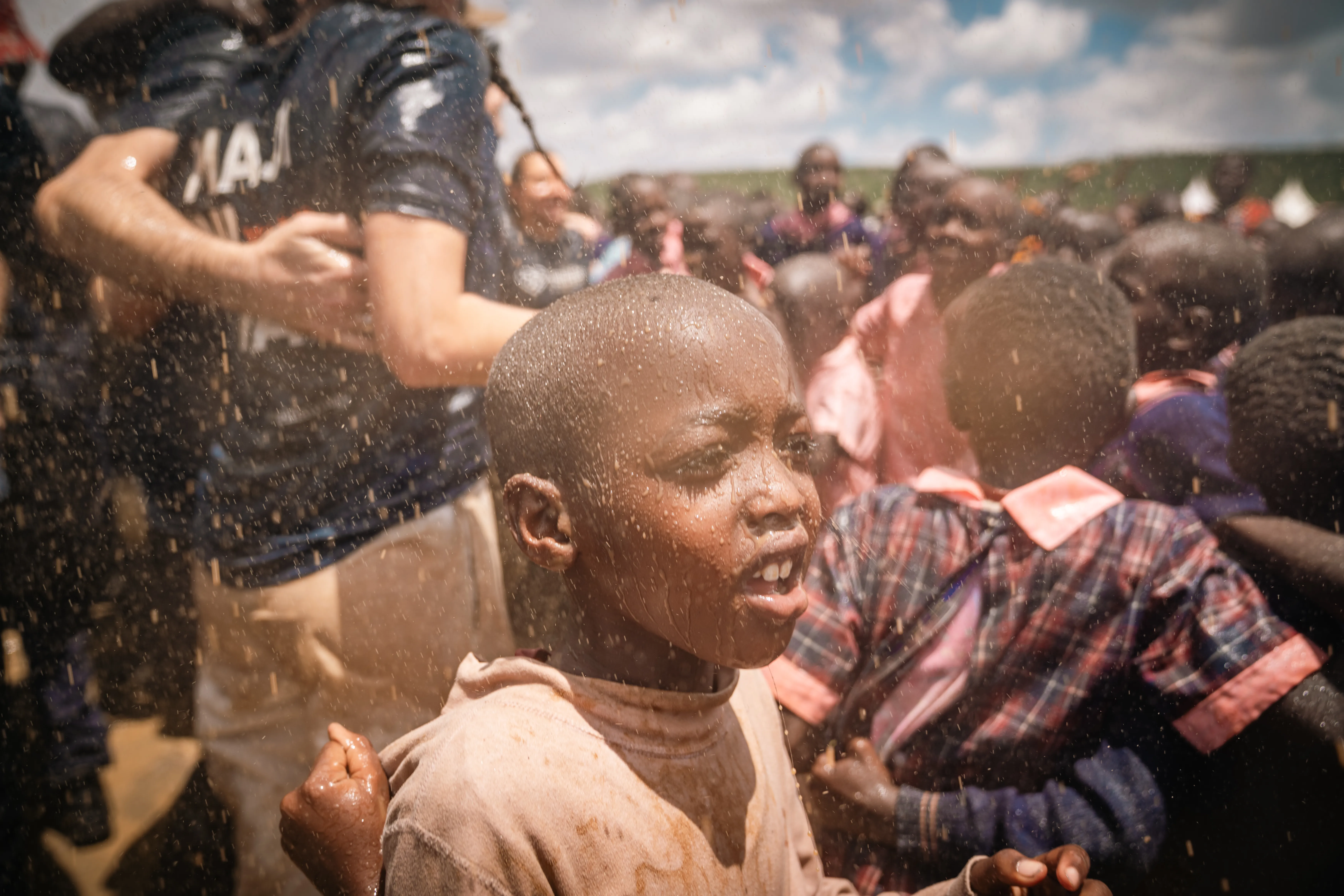 Ein Junge mit nassen Haaren und verschmutzter Haut blickt nach oben, während er von Wasser bespritzt wird. Im Hintergrund sind weitere Menschen zu sehen, die ebenfalls Wasser erhalten.