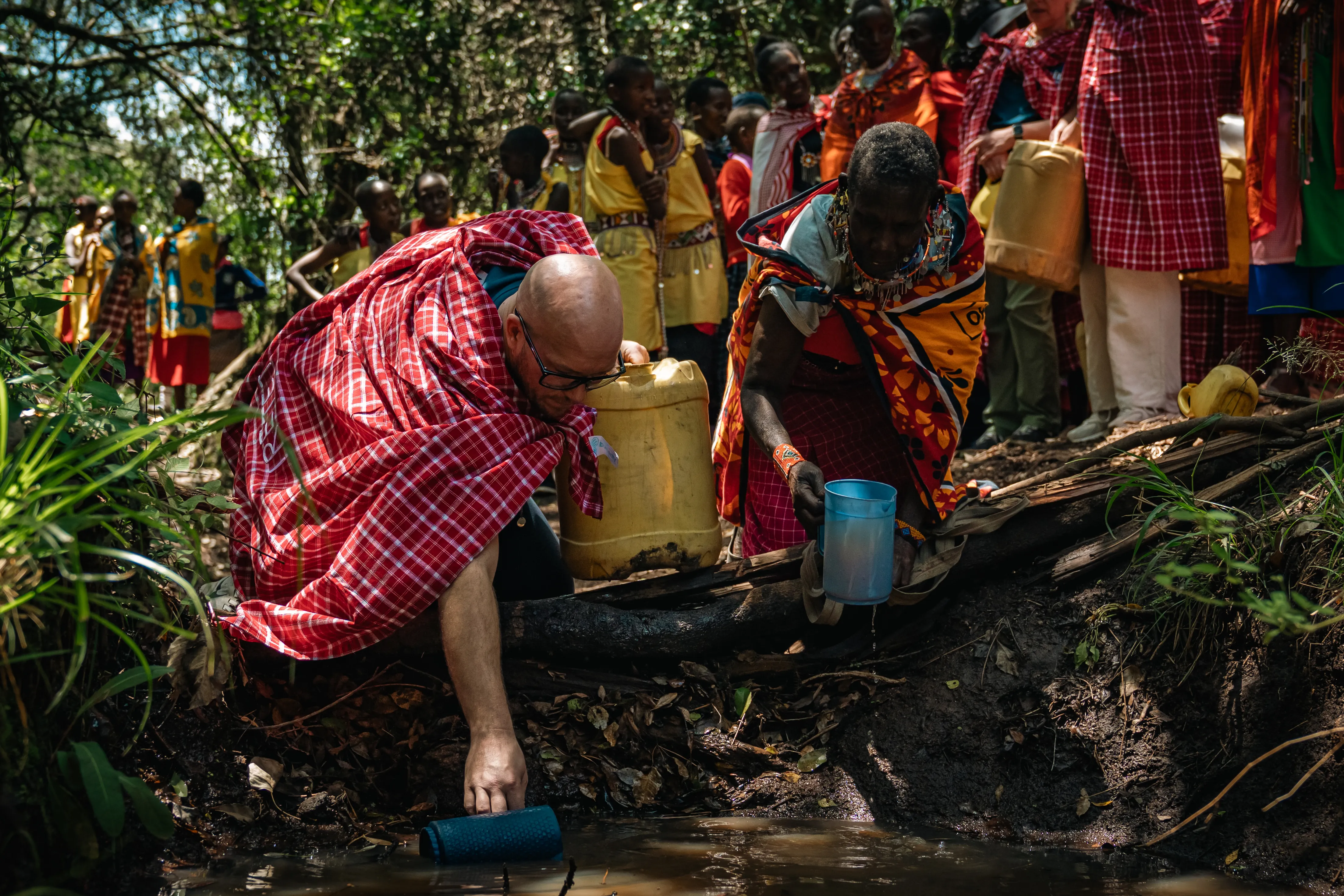 Ein Mann in rot-kariertem Hemd füllt Wasser aus einem kleinen Bach in einen Kanister, während eine Frau mit traditioneller Kleidung daneben steht. Im Hintergrund eine Gruppe von Menschen mit gelben Westen und Kanistern.