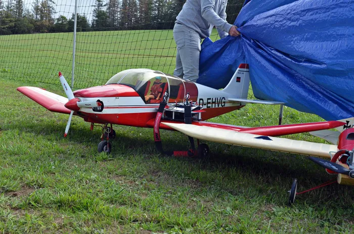Eine Person deckt ein rot-weißes Modellflugzeug auf einer Wiese mit einer blauen Plane ab.