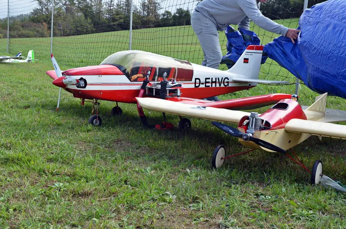Zwei ferngesteuerte Modellflugzeuge auf einer Wiese, im Hintergrund deckt eine Person Ausrüstung mit einer blauen Plane ab.