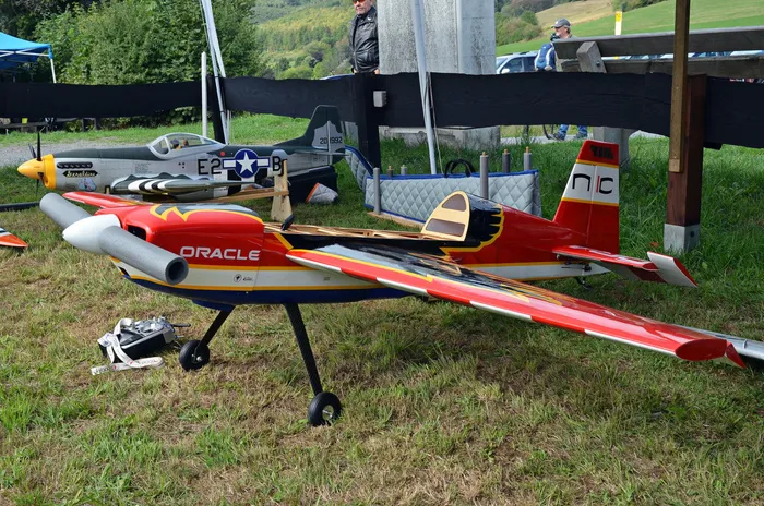 Ein rotes Modellflugzeug mit der Aufschrift „ORACLE“ und ein graues Modellflugzeug im Militärdesign stehen auf einer Wiese.