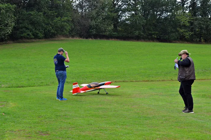 Zwei Männer stehen auf einer Wiese neben einem rot-weißen Modellflugzeug, einer hält eine Fernsteuerung.