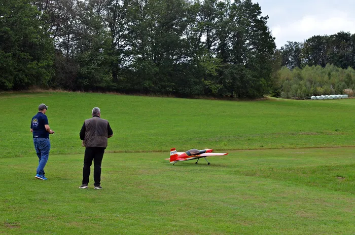Zwei Männer beobachten ein rot-weißes Modellflugzeug auf einer Rasenfläche vor einer Baumkulisse.