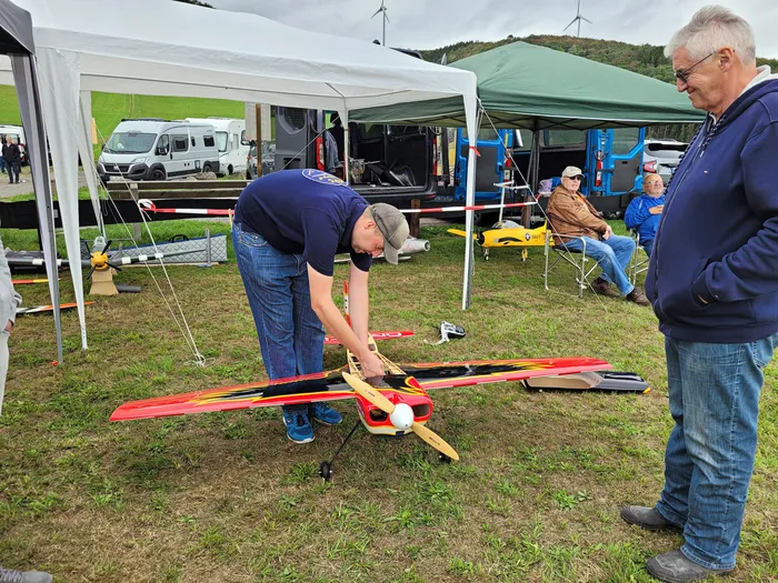 Ein Mann bereitet ein rot-gelbes Modellflugzeug auf einer Wiese vor, während ein anderer Mann daneben steht.