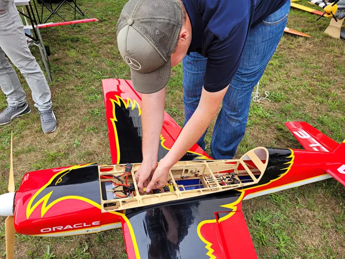 Ein Mann arbeitet an der Technik eines rot-schwarzen Modellflugzeugs mit der Aufschrift „ORACLE“ auf einer Wiese.