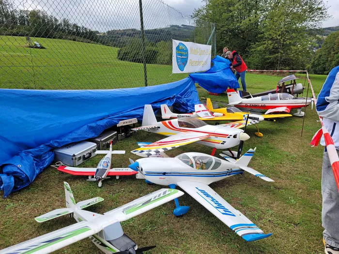 Mehrere bunte Modellflugzeuge liegen auf einer Wiese vor einem Zaun und einer blauen Plane.